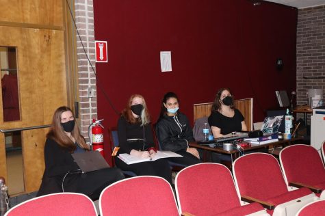 Four people in black and wearing masks sitting and looking at the camera, in the back of the theater.