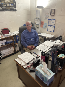 Mr. Tuel sits at his desk. 